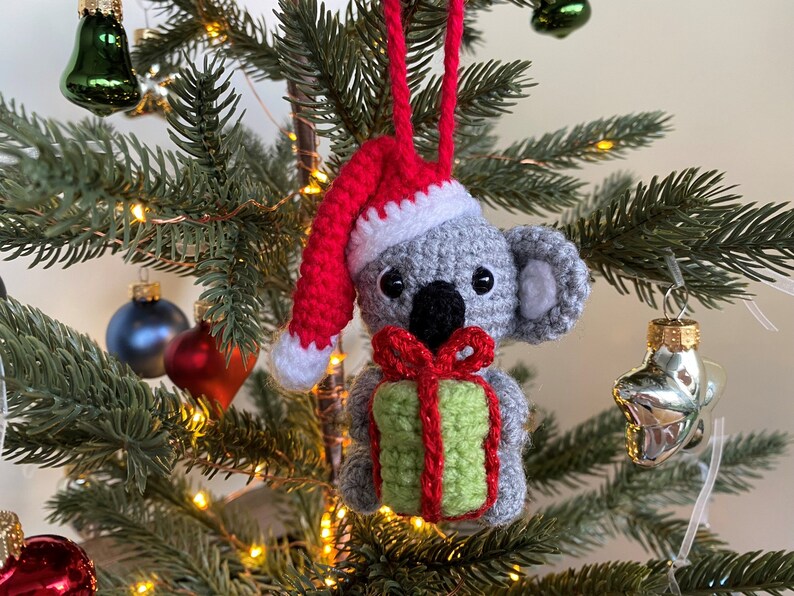 Picture of a crochet Christmas koala. The grey koala is holding a green and red christmas present and is wearing a red Santa hat. The koala is facing forward and hangs on a decorated Christmas tree as an ornament