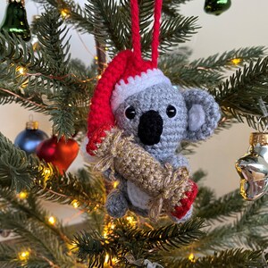 Picture of a crochet Christmas koala. The grey koala is holding a red and gold Christmas cracker and is wearing a red Santa hat. The koala is facing forward and hangs on a decorated Christmas tree as an ornament
