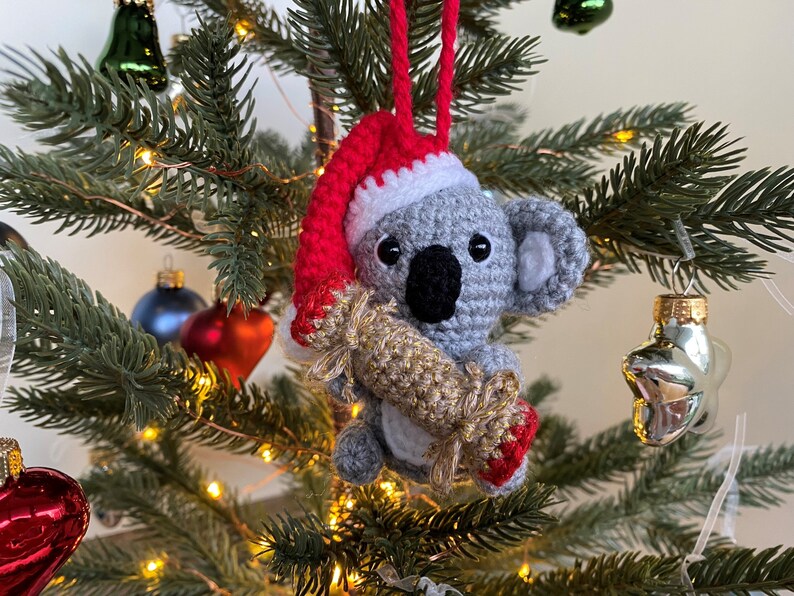 Picture of a crochet Christmas koala. The grey koala is holding a red and gold Christmas cracker and is wearing a red Santa hat. The koala is facing forward and hangs on a decorated Christmas tree as an ornament