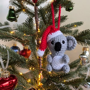 Picture of a crochet Christmas koala. The grey koala is wearing a red Santa hat. The koala is facing forward and hangs on a decorated Christmas tree as an ornament.