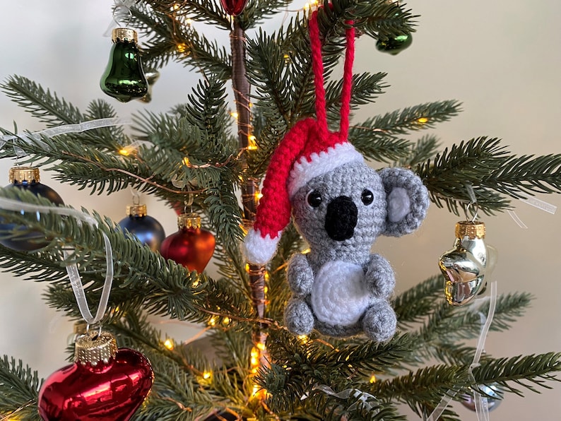 Picture of a crochet Christmas koala. The grey koala is wearing a red Santa hat. The koala is facing forward and hangs on a decorated Christmas tree as an ornament.