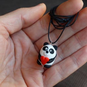 K&ouml;nnte beinhalten: Eine Halskette mit einem Anh&auml;nger in Form eines schwarz-wei&szlig;en Pandas, der ein rotes Herz h&auml;lt. Der Anh&auml;nger ist aus Polymer Clay gefertigt und an einer schwarzen Schnur befestigt.