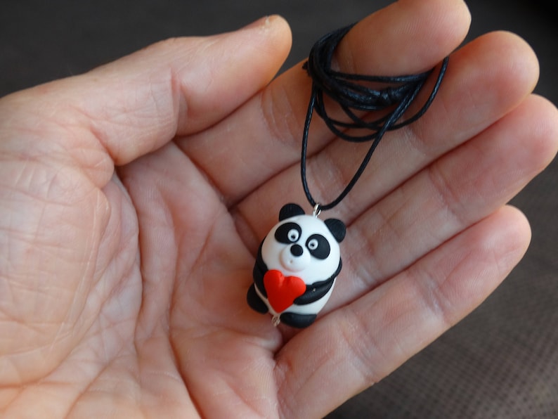 K&ouml;nnte beinhalten: Eine Halskette mit einem Anh&auml;nger in Form eines schwarz-wei&szlig;en Pandas, der ein rotes Herz h&auml;lt. Der Anh&auml;nger ist aus Polymer Clay gefertigt und an einer schwarzen Schnur befestigt.