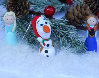 Può includere: Decorazioni natalizie fatte a mano con personaggi di un popolare film d'animazione. Gli ornamenti includono Elsa in un vestito blu, Olaf il pupazzo di neve e Anna in un vestito blu e nero. Sono ambientati su uno sfondo di aghi di pino e neve.