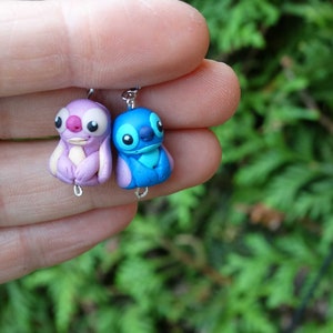 May include: A pair of handmade polymer clay earrings featuring Stitch and Angel from the Disney movie Lilo & Stitch. The earrings are blue and purple and have silver hooks.