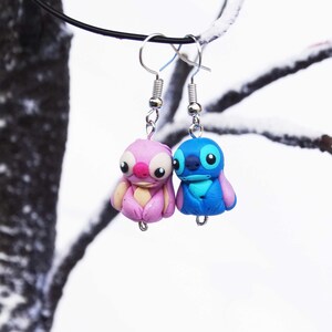 May include: A pair of dangle earrings featuring blue and pink cartoon characters. The characters are stylized and have large eyes. The earrings are hanging from silver hooks.
