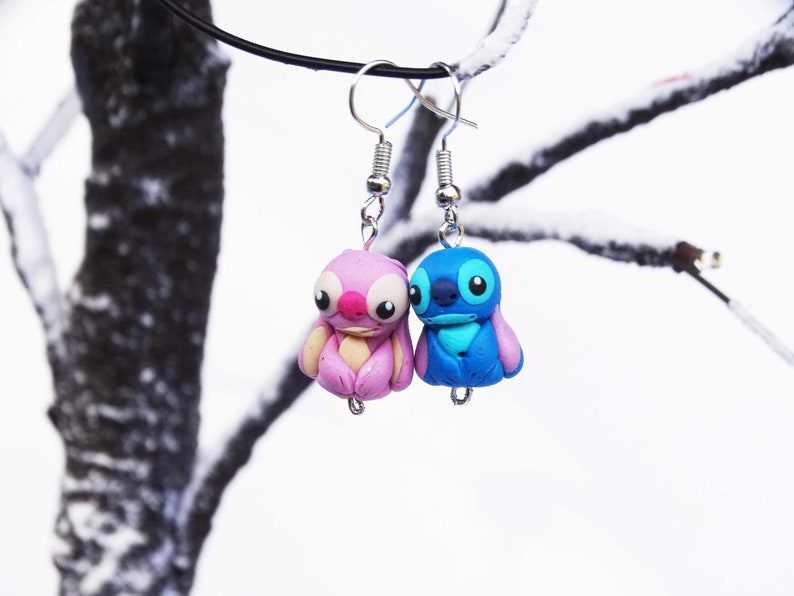 May include: A pair of dangle earrings featuring blue and pink cartoon characters. The characters are stylized and have large eyes. The earrings are hanging from silver hooks.
