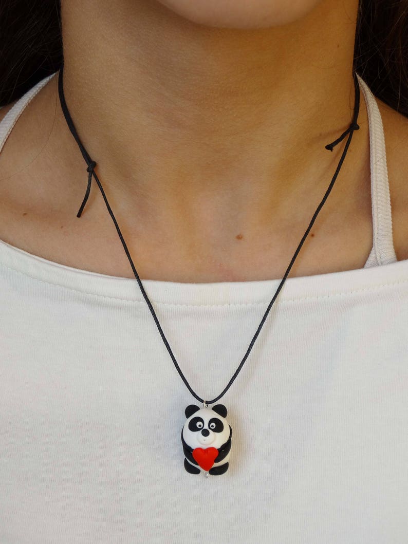 K&ouml;nnte beinhalten: Eine schwarze Schnurkette mit einem wei&szlig;en und schwarzen Pandabanh&auml;nger, der ein rotes Herz h&auml;lt. Der Pandabanh&auml;nger ist aus Polymer Clay.
