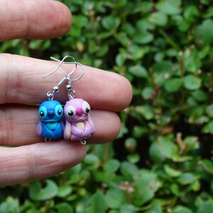 May include: A pair of dangle earrings featuring blue and purple Stitch and Angel characters from the Disney movie Lilo & Stitch. The earrings are made of polymer clay and have silver hooks.
