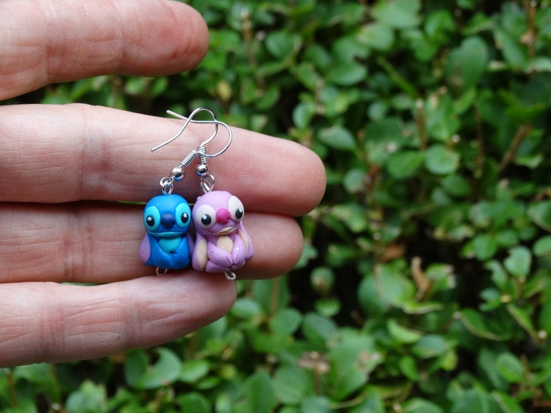 May include: A pair of dangle earrings featuring blue and purple Stitch and Angel characters from the Disney movie Lilo & Stitch. The earrings are made of polymer clay and have silver hooks.