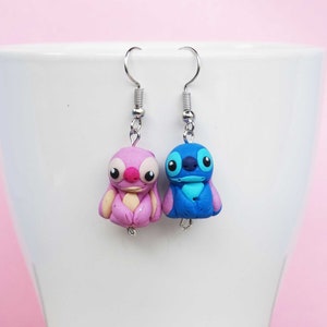 May include: A pair of dangle earrings featuring a pink and blue Stitch character from the Disney movie Lilo & Stitch. The earrings are made of polymer clay and have silver hooks.