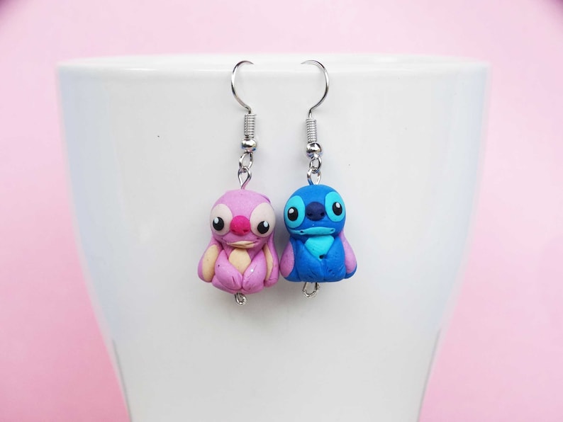 May include: A pair of dangle earrings featuring a pink and blue Stitch character from the Disney movie Lilo & Stitch. The earrings are made of polymer clay and have silver hooks.