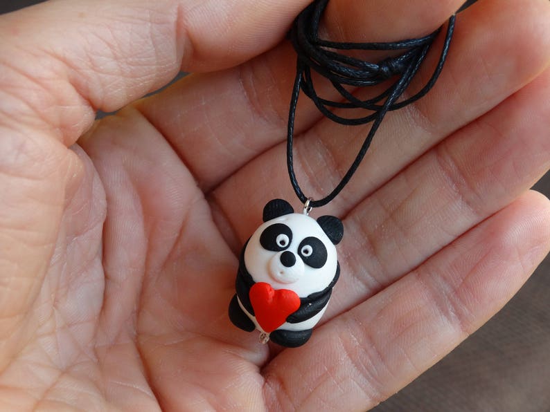 K&ouml;nnte beinhalten: Eine schwarze Schnurkette mit einem Panda-Anh&auml;nger, der ein rotes Herz h&auml;lt. Der Panda ist aus wei&szlig;em und schwarzem Fimo gefertigt.