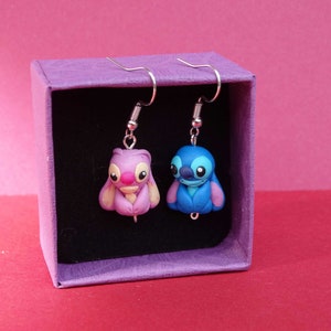 May include: A pair of dangle earrings featuring blue and pink cartoon characters in a purple box. The characters are Stitch and Angel from the Disney movie Lilo & Stitch.