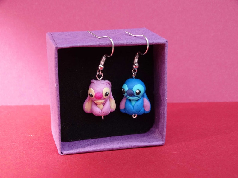 May include: A pair of dangle earrings featuring blue and pink cartoon characters in a purple box. The characters are Stitch and Angel from the Disney movie Lilo & Stitch.