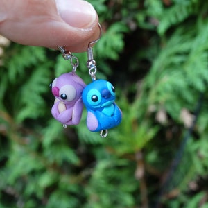 May include: A pair of dangle earrings featuring Stitch and Angel from the Disney movie Lilo & Stitch. The earrings are made of polymer clay and are painted in blue, purple, and pink.