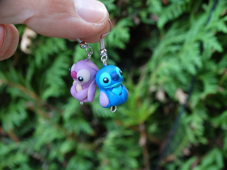 May include: A pair of dangle earrings featuring Stitch and Angel from the Disney movie Lilo & Stitch. The earrings are made of polymer clay and are painted in blue, purple, and pink.