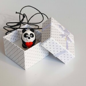 K&ouml;nnte beinhalten: Eine wei&szlig;e Geschenkbox mit Punkten und einem wei&szlig;en Band, ge&ouml;ffnet, um einen schwarz-wei&szlig;en Pandabanh&auml;nger zu enth&uuml;llen, der ein rotes Herz h&auml;lt. Der Anh&auml;nger ist an einer schwarzen Schnur befestigt.