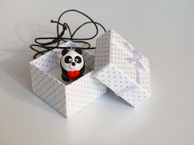K&ouml;nnte beinhalten: Eine wei&szlig;e Geschenkbox mit Punkten und einem wei&szlig;en Band, ge&ouml;ffnet, um einen schwarz-wei&szlig;en Pandabanh&auml;nger zu enth&uuml;llen, der ein rotes Herz h&auml;lt. Der Anh&auml;nger ist an einer schwarzen Schnur befestigt.