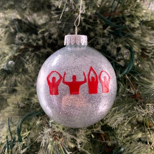 May include: A silver glitter Christmas ornament with three red stick figures in a raised hand pose.