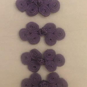 May include: Five sets of purple frog closures, each composed of two flower-shaped knots connected by a small knot. The closures are made of a tightly wound, textured material, and are arranged vertically on a light background.
