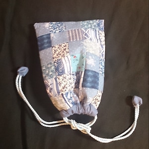 Puede incluir: Una bolsa de cordón azul hecha de tela de patchwork con un cordón blanco. La bolsa está cerrada y tiene una variedad de patrones azules, blancos y crema.