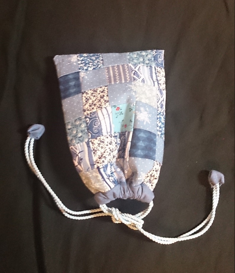 May include: A blue drawstring bag made of patchwork fabric with a white cord. The bag is tied closed and has a variety of blue, white, and cream patterns.
