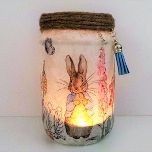 Pode incluir: Um frasco de vidro branco com uma tampa de corda marrom e um tassel azul. O frasco é decorado com uma imagem impressa do Peter Rabbit e flores. Uma pequena vela está dentro do frasco.
