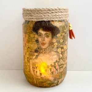 May include: A decorative glass jar featuring a portrait of a woman in a gold and brown design. The jar has a rope-wrapped rim and a small, orange tassel. The jar is lit from within, creating a warm glow.