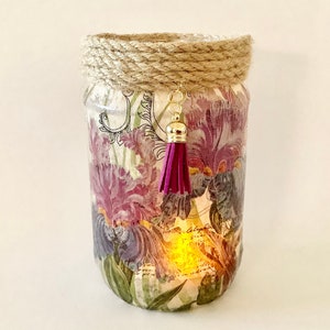 May include: A glass jar candle holder with a floral design featuring purple and white irises. The jar is decorated with a brown rope around the top and a purple tassel hanging from the lid.