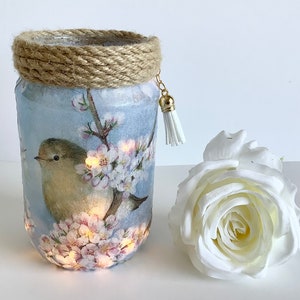 Op de afbeelding: Een glazen pot met een blauwe achtergrond en een witte vogel met roze bloemen. De pot heeft een bruin touw om de bovenkant en een gouden kwast die er aan hangt.