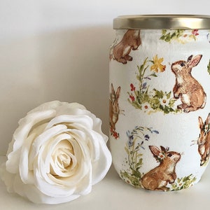 以下が含まれることがあります： 金色の蓋が付いた白い瓶で、茶色のウサギと花の模様で飾られています。瓶の隣には大きな白いバラがあります。瓶は装飾品です。