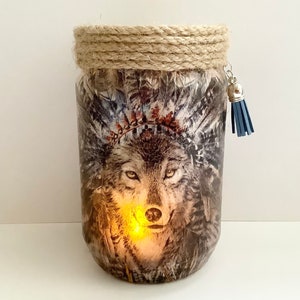 Op de afbeelding: Een glazen pot met een wolvenkop en een hoofdtooi van veren. De pot is versierd met een bruin touw rond de bovenkant en een blauwe kwast.