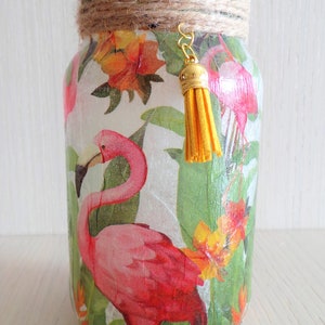 Könnte beinhalten: Ein Glasgefäß, verziert mit einem tropischen Flamingo- und Blumendesign. Das Gefäß hat einen braunen Kordeldeckel und einen goldenen Quastenanhänger.