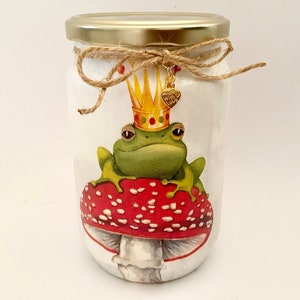 Peut inclure: Un bocal en verre décoré d'une grenouille verte portant une couronne dorée assise sur un champignon rouge et blanc. Le bocal est orné d'un nœud en ficelle brune et d'un charme doré avec les mots "Faites un vœu" dessus.
