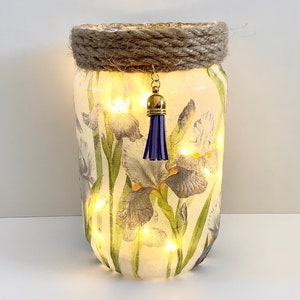 May include: A glass jar with a white background and a floral design of purple and white irises. The jar has a brown rope around the top and a purple tassel hanging from the lid. The jar is lit from within with white lights.