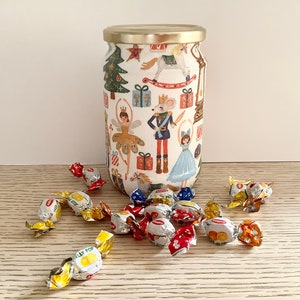 Può includere: Un barattolo di vetro con coperchio dorato, decorato con un design festivo a tema Schiaccianoci. Il barattolo è pieno di caramelle incartate. Il design include un albero di Natale, una ballerina e un soldatino.