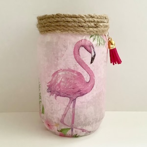 May include: A pink glass jar with a pink flamingo design and a brown twine accent around the top. The jar has a pink tassel hanging from the top.