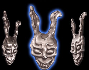 May include: A silver 3D enamel pin featuring the iconic Frank the Bunny mask from the film Donnie Darko. The pin is a detailed replica of the mask, with a menacing grin and large, rabbit-like ears.