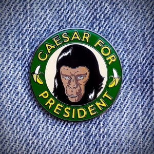 Może przedstawiać: Zielono-ż&oacute;łta szpilka emaliowana z czarno-białym obrazkiem Cezara z filmu "Planeta Małp". Szpilka zawiera napis "Caesar for President" z dwoma ż&oacute;łtymi bananami po obu stronach tekstu.