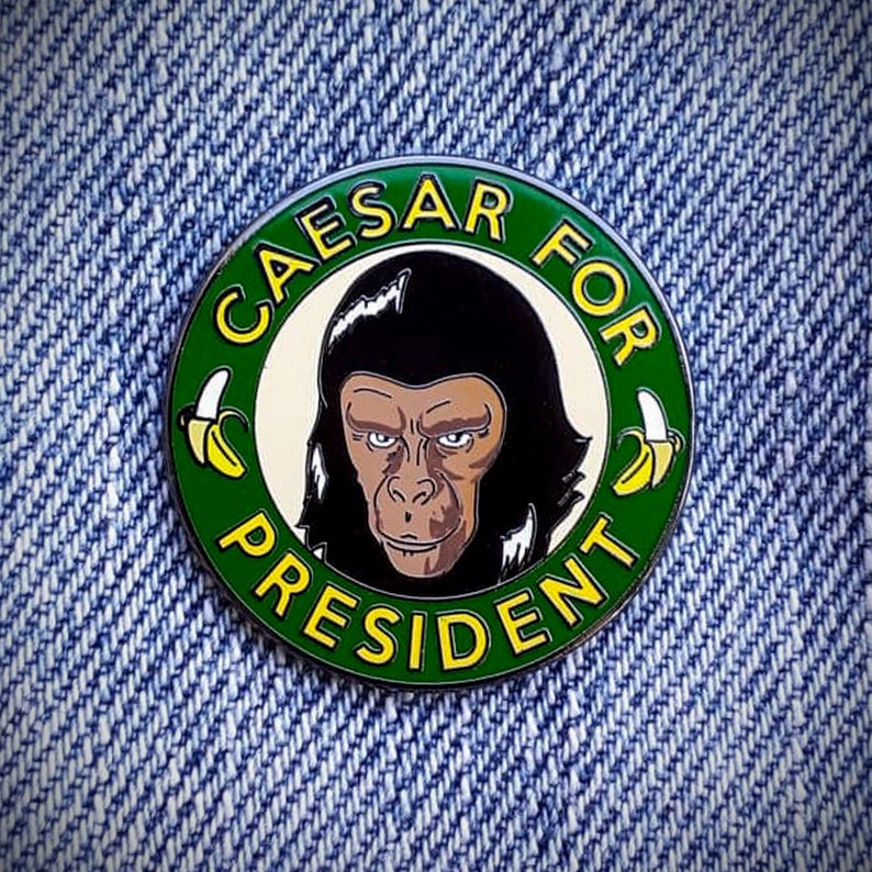 Może przedstawiać: Zielono-ż&oacute;łta szpilka emaliowana z czarno-białym obrazkiem Cezara z filmu "Planeta Małp". Szpilka zawiera napis "Caesar for President" z dwoma ż&oacute;łtymi bananami po obu stronach tekstu.