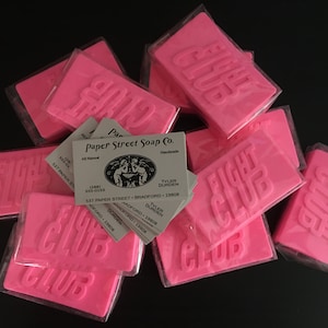 May include: A stack of pink soap bars with the words "Fight Club" embossed on them. The soap bars are wrapped in clear plastic and are sitting on a black surface. There are also several business cards for "Paper Street Soap Co." visible in the image.