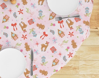 Puede incluir: Un mantel rosa con un estampado navideño festivo. El diseño incluye hombrecitos de jengibre, ciervos, adornos y cajas de regalo. Dos platos blancos, un tenedor y un cuchillo están puestos en la mesa para una comida festiva.