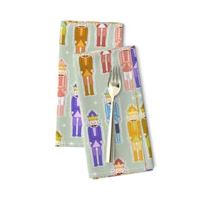 Op de afbeelding: Twee groene stoffen servetten met een herhalend patroon van kleurrijke notenkrakersoldaten. De notenkrakers dragen hoeden en uniformen in verschillende kleuren. De achtergrond is lichtgroen met witte sterren.