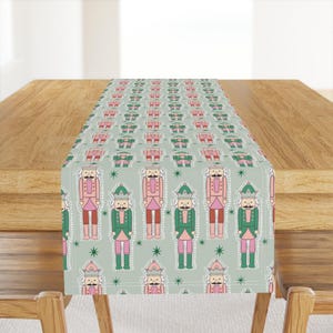 May include: A green and pink table runner with a repeating pattern of nutcracker soldiers. The nutcrackers are wearing red and pink hats and have green and white uniforms.
