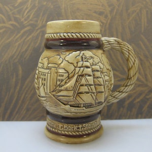 May include: A brown ceramic beer stein with a rope-like handle. The stein features a detailed relief design of a ship sailing past a city skyline. The text "antine T. Bork T. Stauson" is visible on the base.
