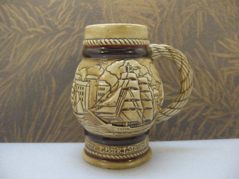 May include: A brown ceramic beer stein with a rope-like handle. The stein features a detailed relief design of a ship sailing past a city skyline. The text "antine T. Bork T. Stauson" is visible on the base.