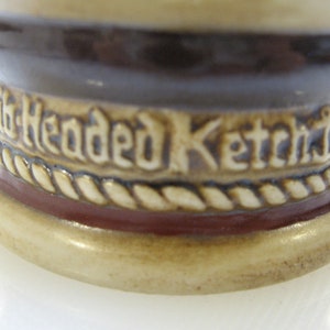 May include: Close-up of the bottom of a ceramic stein with a brown and white rope design and the words "Six Headed Ketch" in white lettering.