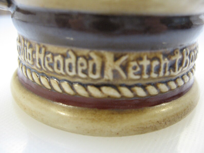 May include: Close-up of the bottom of a ceramic stein with a brown and white rope design and the words "Six Headed Ketch" in white lettering.