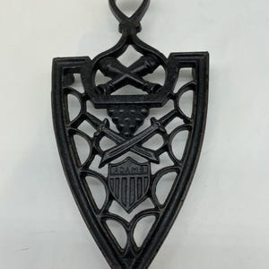 May include: Black cast iron trivet with a shield shape. The trivet features crossed cannons, swords, and a shield with the word "ADAMS". The handle has a decorative floral design.